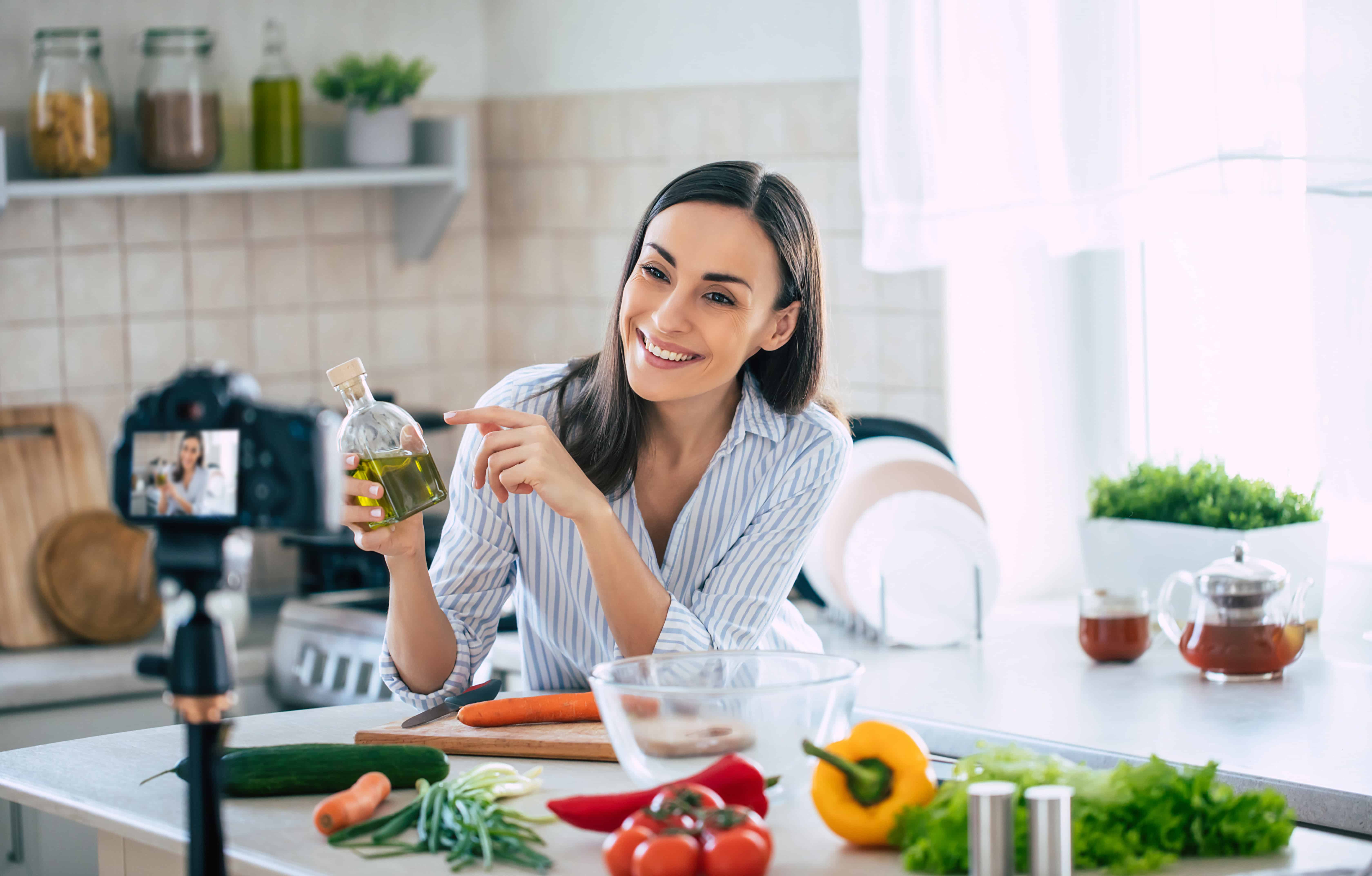 Professional beautiful happy young woman is blogging for her kitchen channel about healthy living in the kitchen of her home and looking on camera on a tripod
